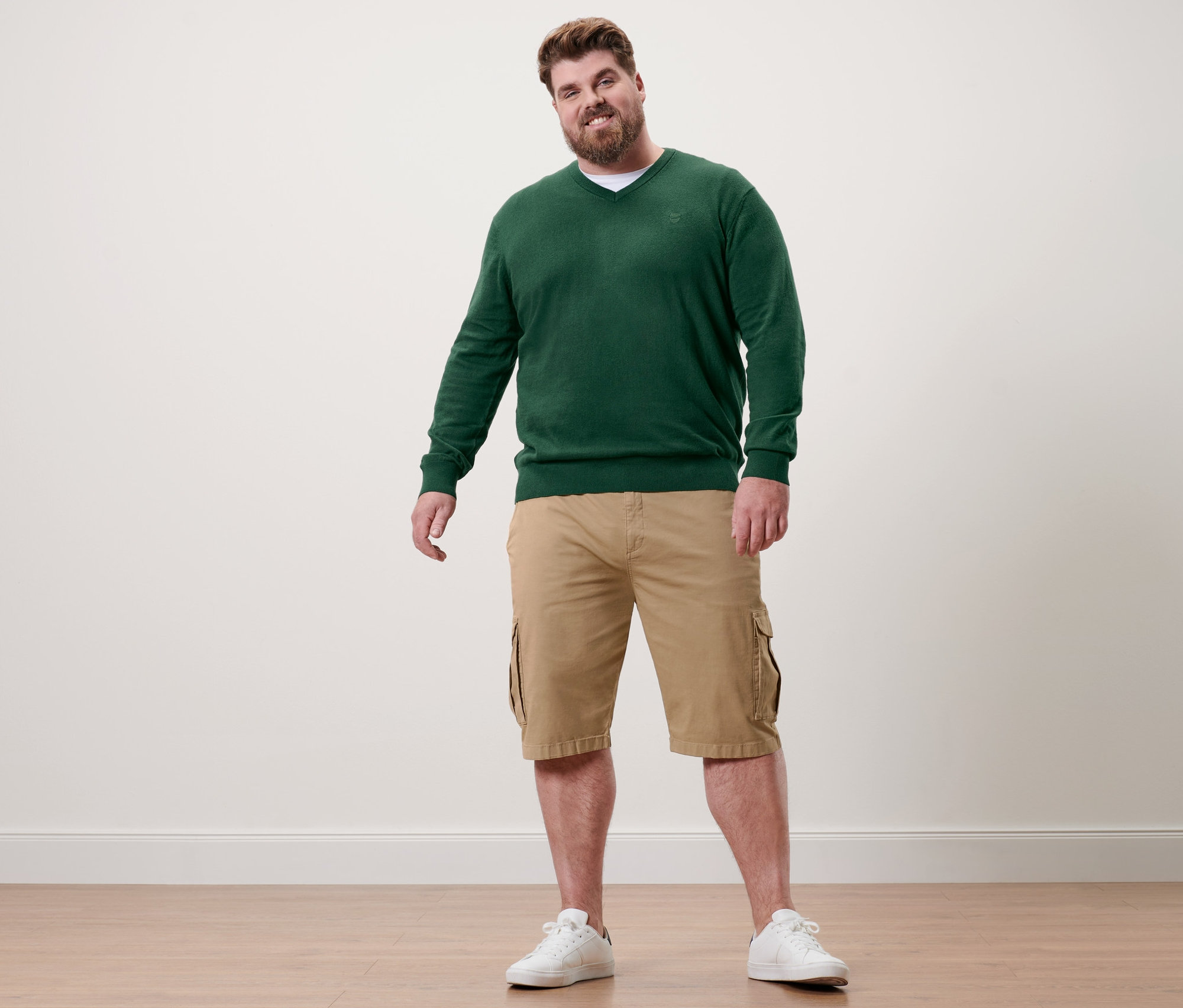 Mann steht und trägt einen dunkelgrünen Pullover mit V-Ausschnitt, Khaki-Shorts und weiße Turnschuhe.