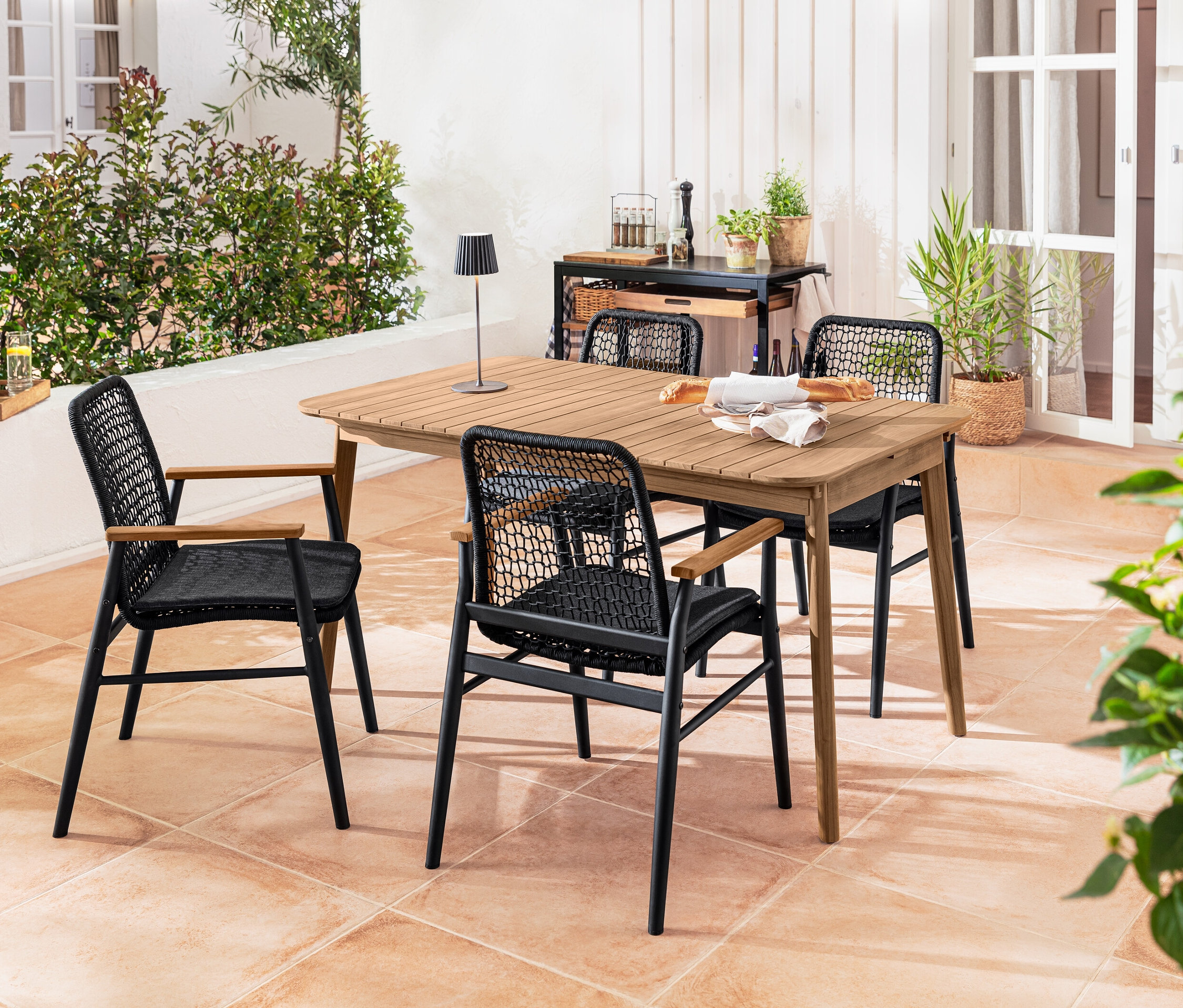 Vier Dining-Stühle mit Teak-Armlehnen und Textilgeflecht stehen um einen Teak-Ausziehtisch. Auf dem Tisch steht eine schwarze LED-Akku-Outdoor-Tischleuchte.
