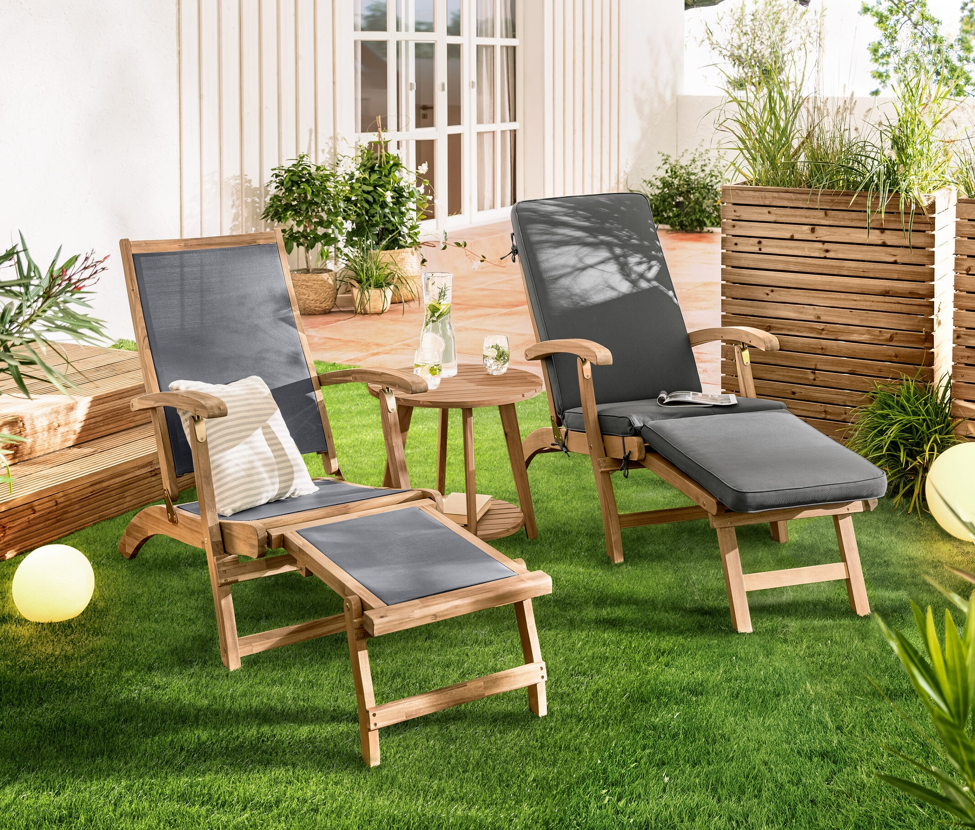 Zwei Deckchairs »Lenja« mit ausziehbarer Fußlehne stehen nebeneinander auf einer Wiese. Dazwischen steht ein Beistelltisch mit 2 Ebenen »Lenja«. Im Hintergrund sind Solar-Kugelleuchten, Sandoptik, ca. 20 cm und Solar-Kugelleuchten, Sandoptik, ca. 30 cm zu sehen.