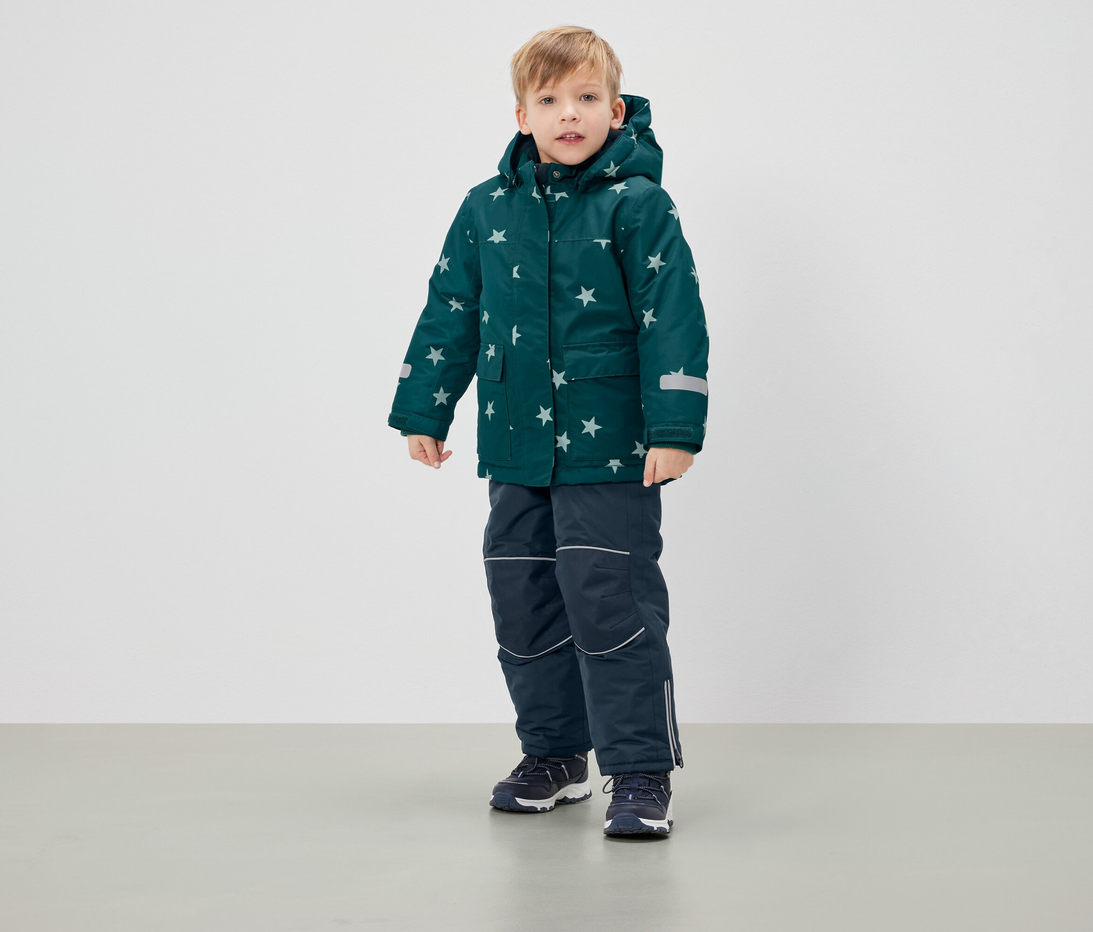 Junge steht in grüner Winterjacke mit Sternen und dunkelblauer Hose.