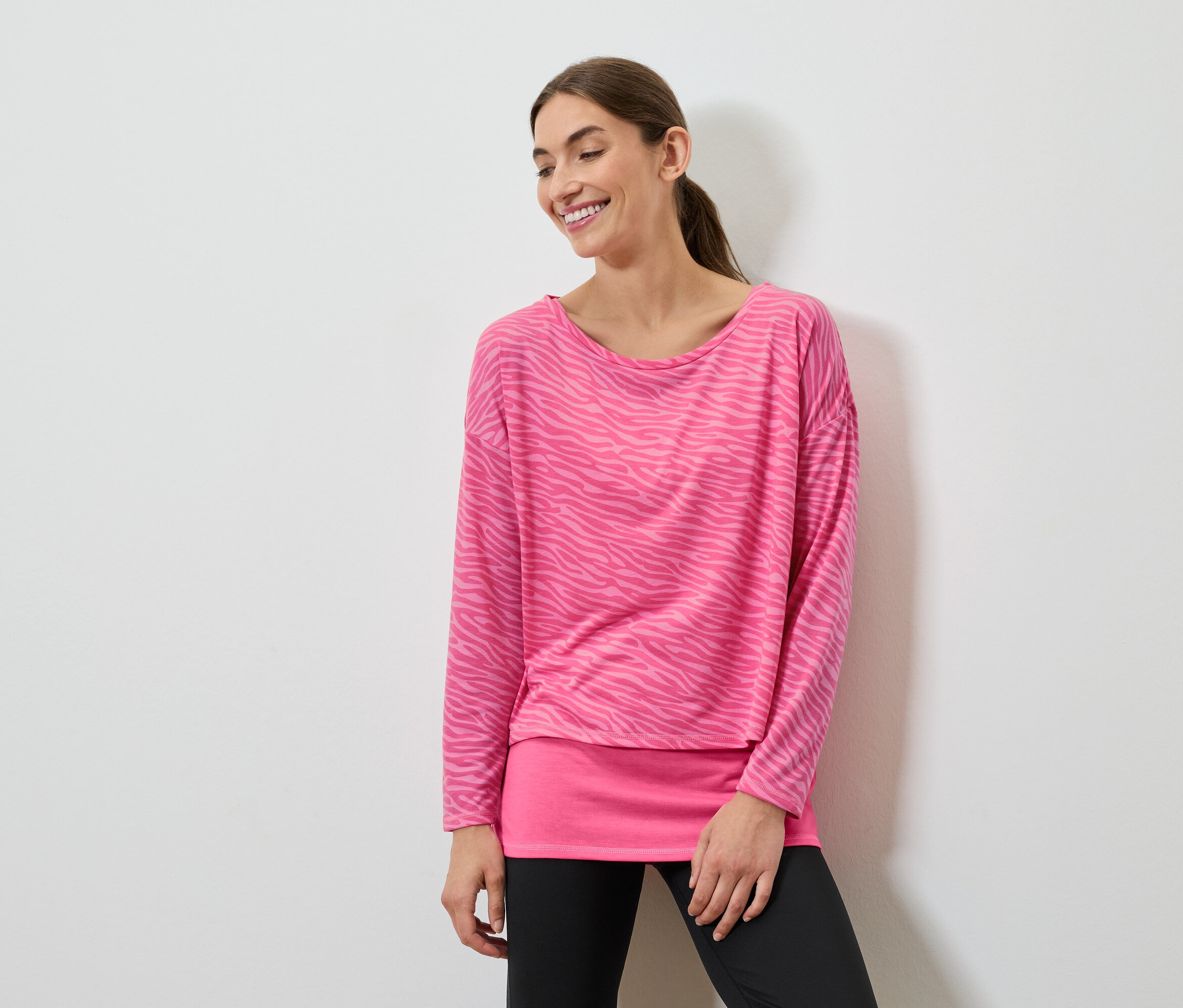 Lächelnde Frau posiert in einem pinkfarbenen Langarmshirt und schwarzen Leggings.