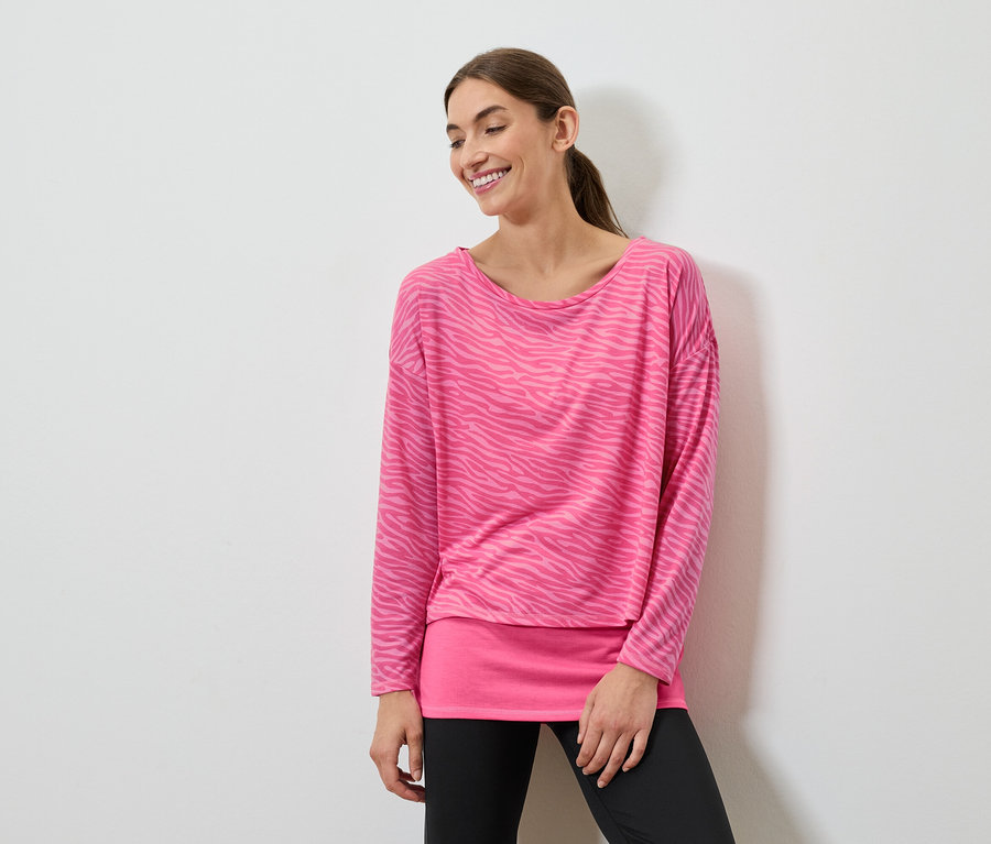 Lächelnde Frau posiert in einem pinkfarbenen Langarmshirt und schwarzen Leggings.