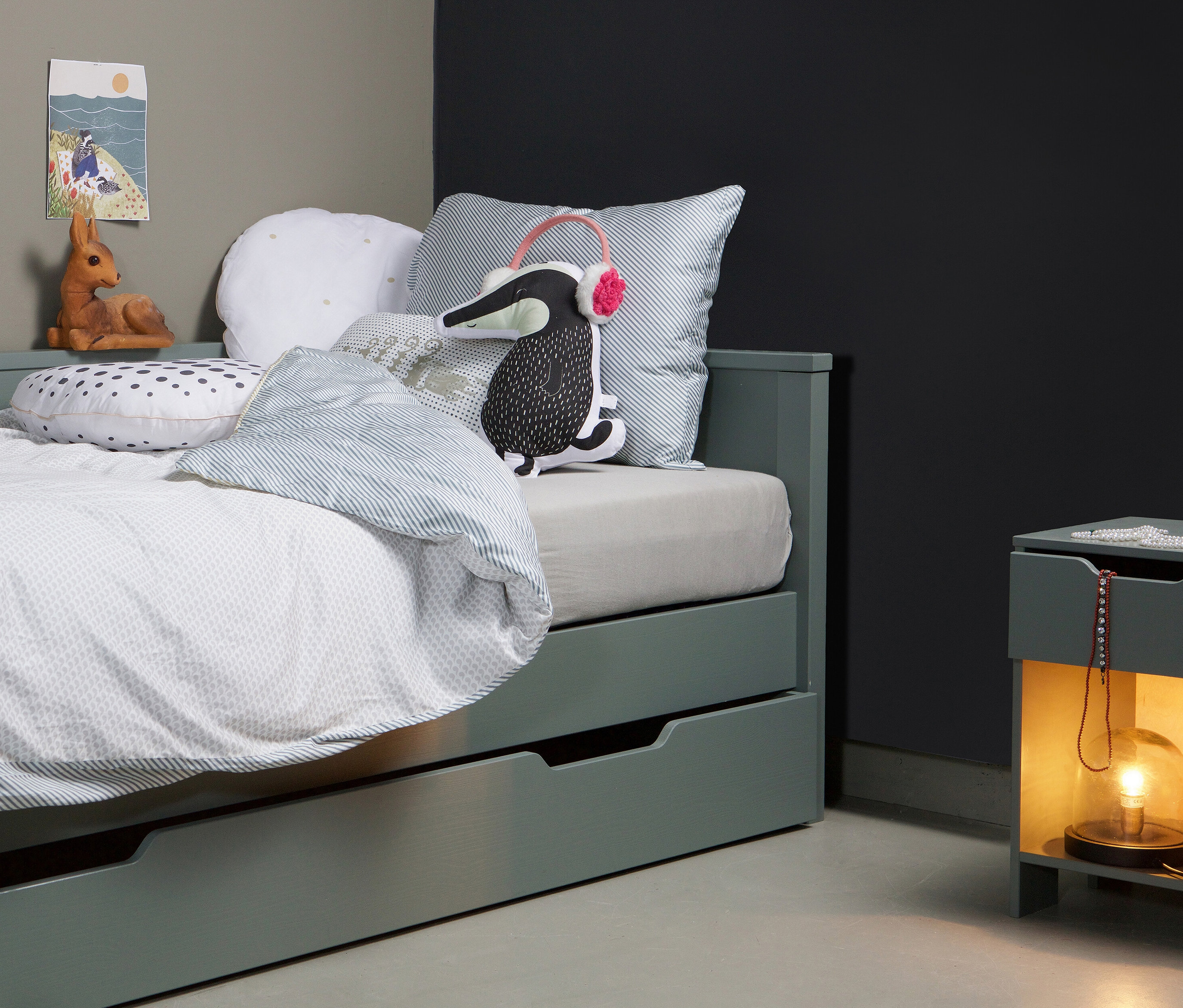 Grünes Bett mit Bettwäsche und Kissen und ein grüner Nachttisch mit Lampe. Unter dem Bett befindet sich ein grüner Woood Bettkasten »Nikki«.