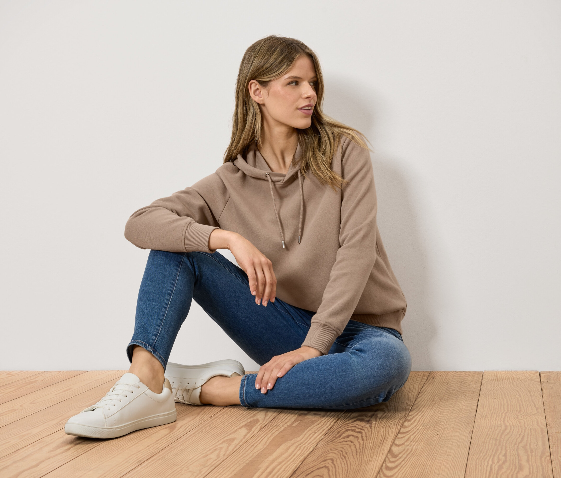 Eine Frau sitzt auf einem Holzboden und trägt einen braunen Kapuzenpullover, blaue Jeans und weiße Turnschuhe.