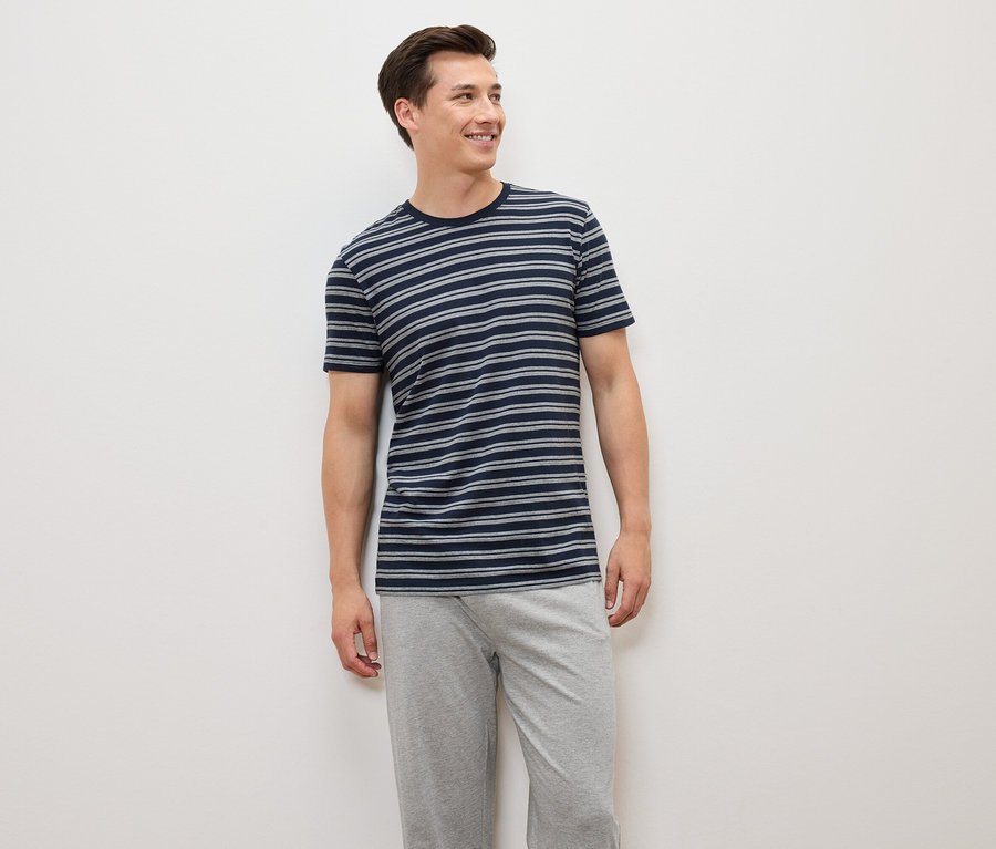 Ein Mann steht und trägt ein blau-grau gestreiftes T-Shirt und graue Hosen.