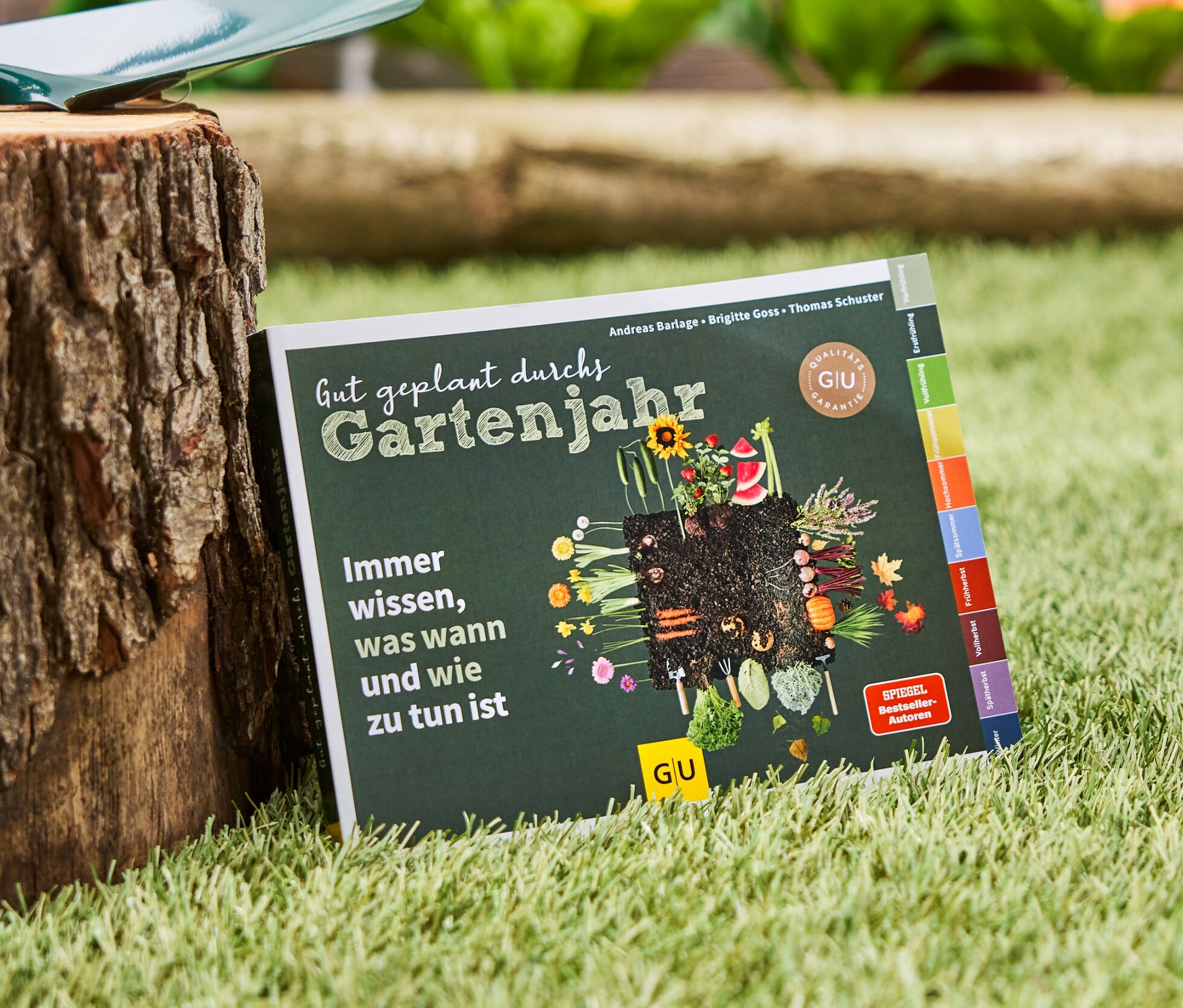 Das Buch »Gut geplant durchs Gartenjahr« liegt auf dem Rasen neben einem Baumstumpf, auf dem eine Gartenschaufel liegt.