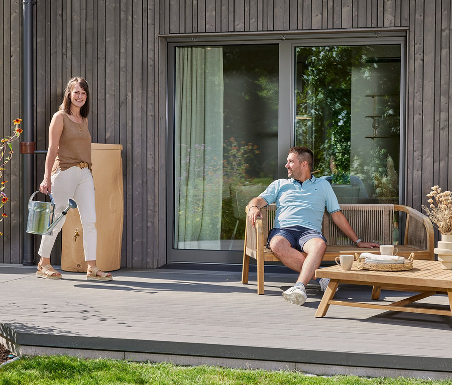 Frau trägt Gießkanne neben Regentonne, Mann sitzt auf Bank.