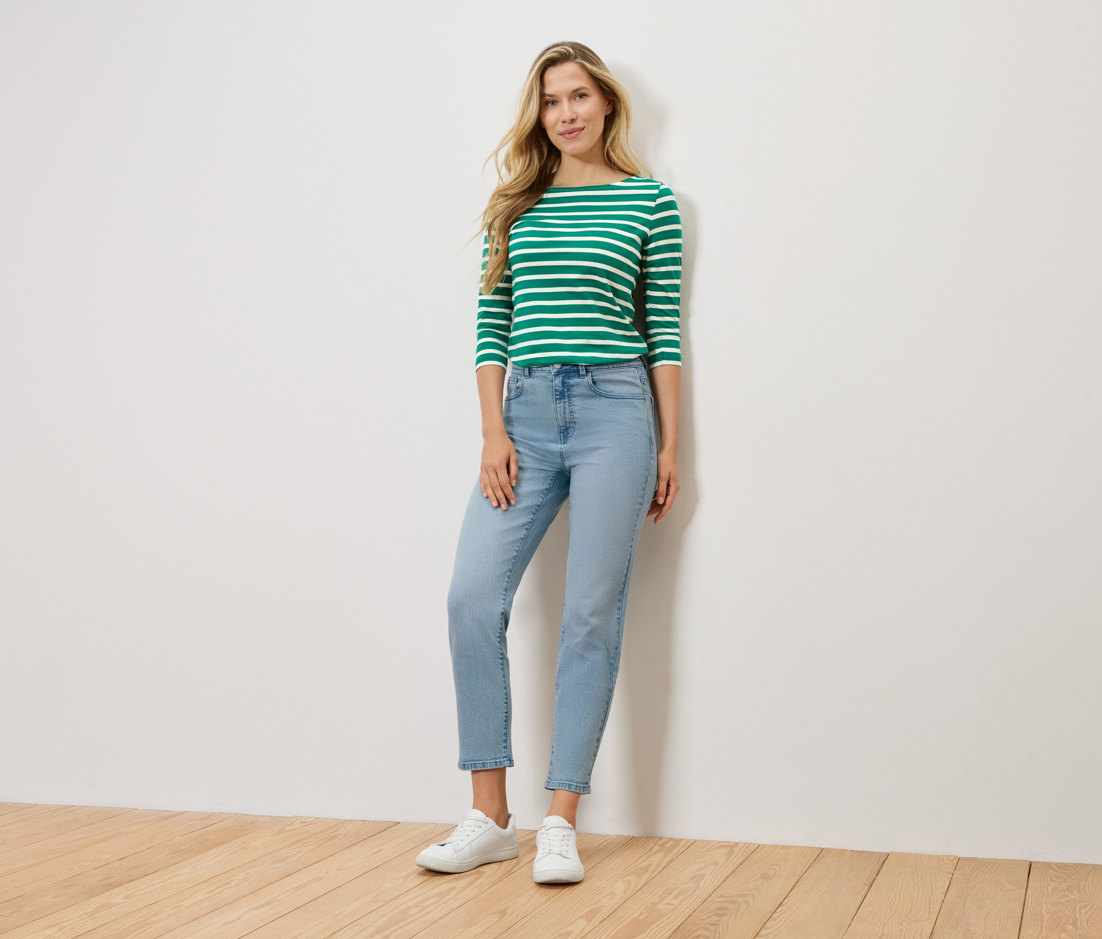 Eine blonde Frau steht vor einer Wand und trägt ein grün-weiß gestreiftes Langarmshirt, hellblaue Jeans und weiße Sneaker.