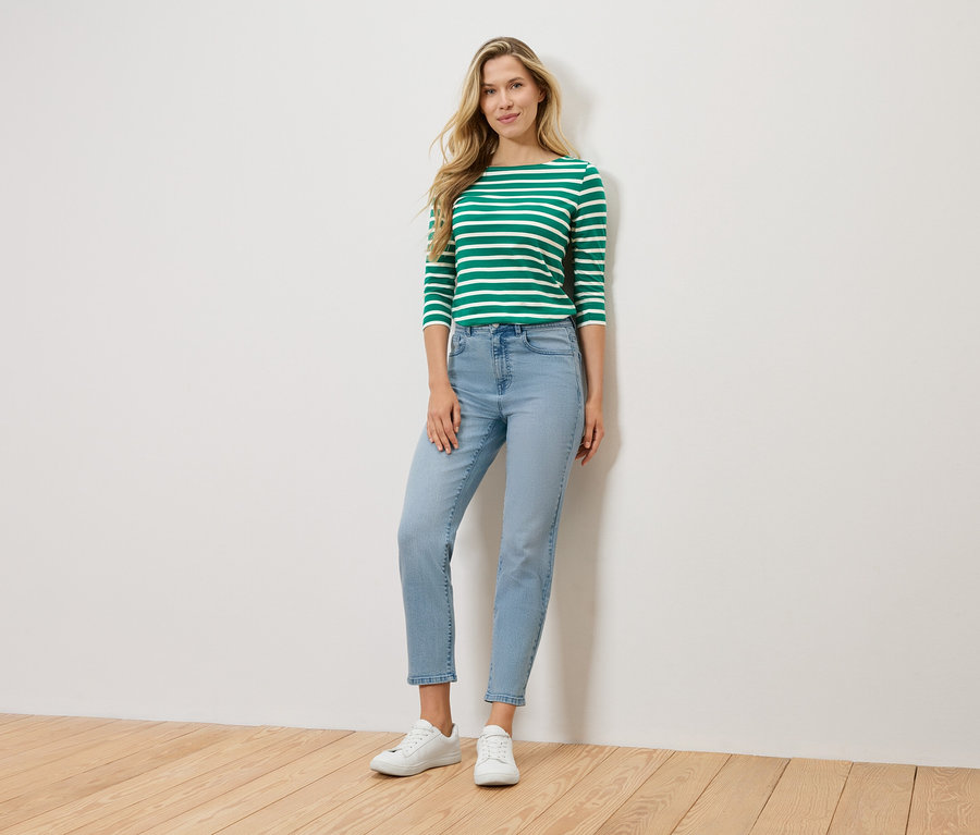 Eine blonde Frau steht vor einer Wand und trägt ein grün-weiß gestreiftes Langarmshirt, hellblaue Jeans und weiße Sneaker.