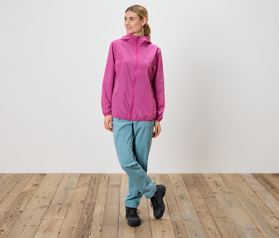 Frau steht mit gekreuzten Beinen, trägt eine pinke Jacke, eine blaue Hose und schwarze Schuhe.