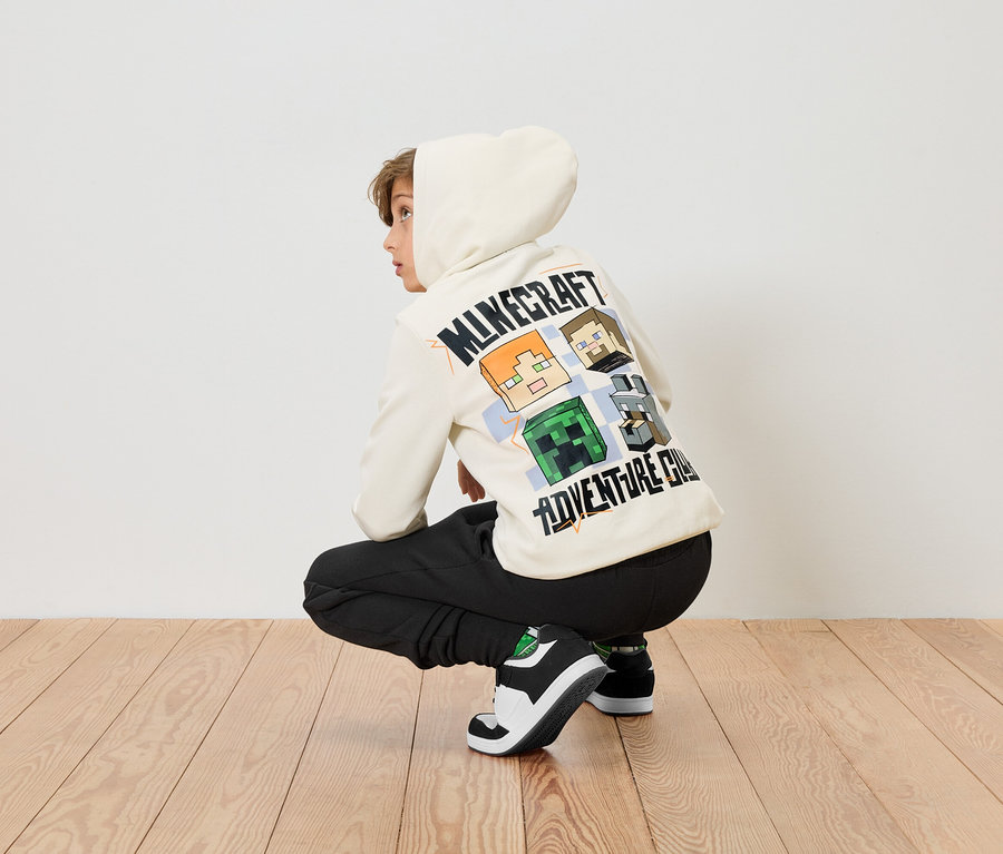 Ein Junge hockt auf einem Holzboden und trägt einen cremefarbenen Minecraft-Hoodie und schwarze Hosen.
