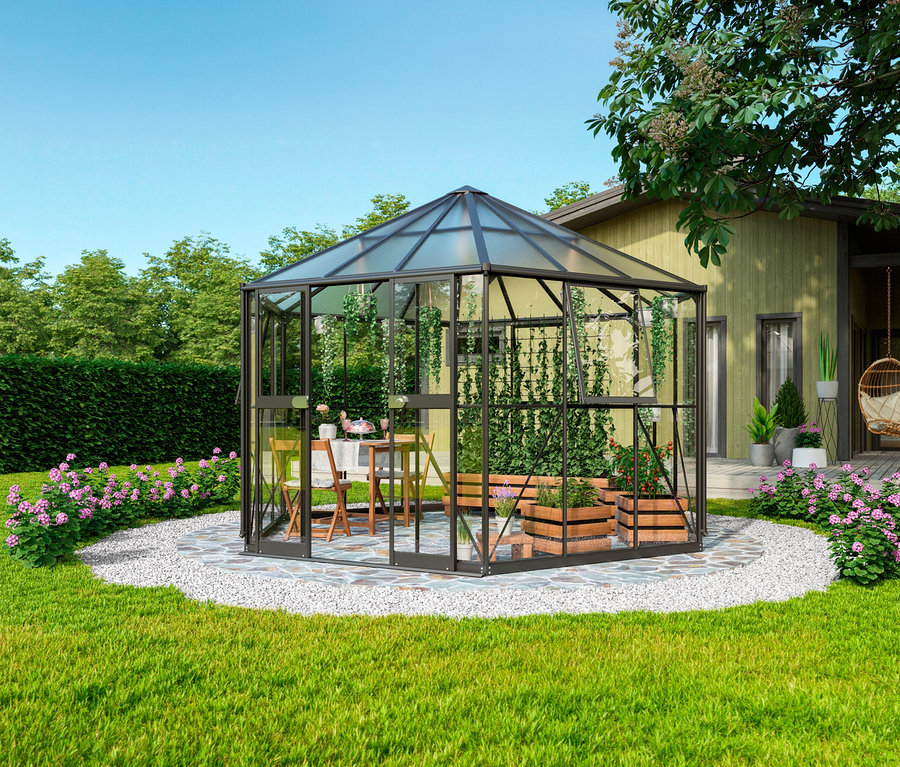 Der schwarze Vitavia Gewächshaus Pavillion 9 m² mit Fundamentrahmen steht auf einem Kiesweg im Garten. Im Inneren stehen ein Tisch und Stühle.