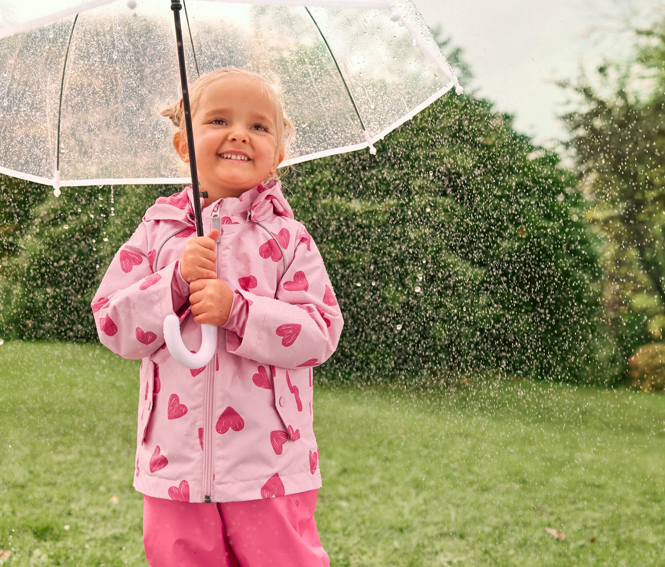 Ein kleines Mädchen mit blonden Haaren lächelt unter einem transparenten Regenschirm im Regen, bekleidet mit einer rosafarbenen Jacke mit Herzmuster.