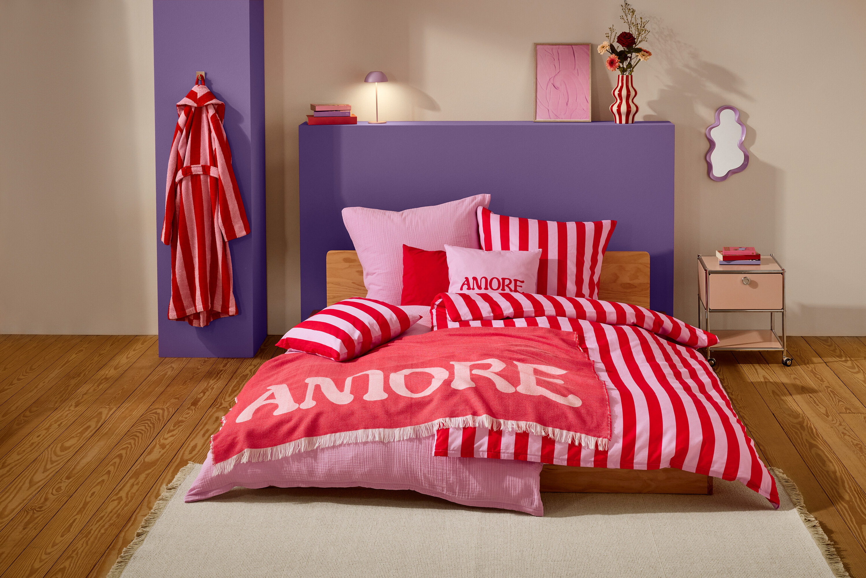 Ein Schlafzimmer mit einem Bett, das mit rot-weiß gestreifter Bettwäsche, rosa Kissen und einer korallenfarbenen Decke mit der Aufschrift "Amore" bedeckt ist. Ein rot-weiß gestreifter Bademantel hängt an einem violetten Pfeiler.