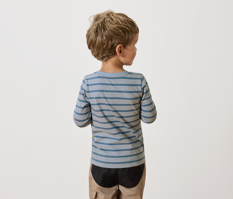 Rückenansicht eines Jungen mit blonden, lockigen Haaren, der ein blau-grau gestreiftes Langarmshirt und beigefarbene Hosen trägt.