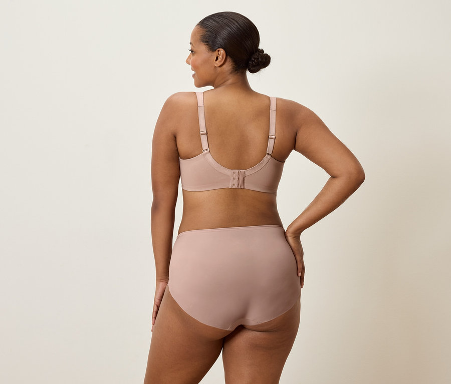 Eine Frau von hinten, die einen hautfarbenen BH und Slip trägt und eine Hand auf die Hüfte legt.