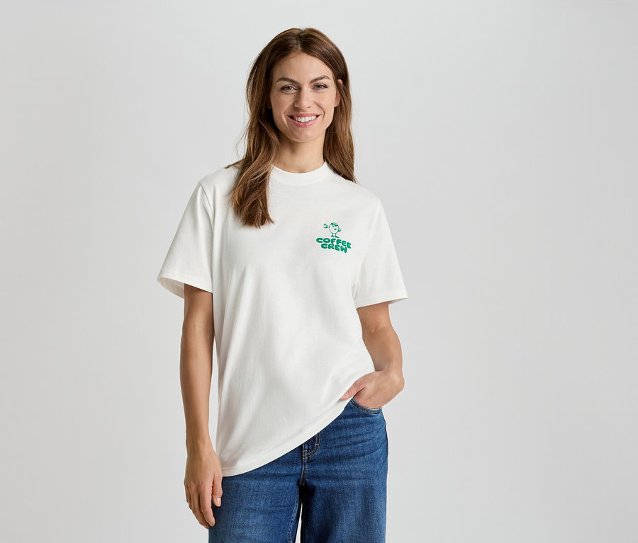 Frau mit weißem T-Shirt mit "Coffee Crew" Aufdruck und blauen Jeans.