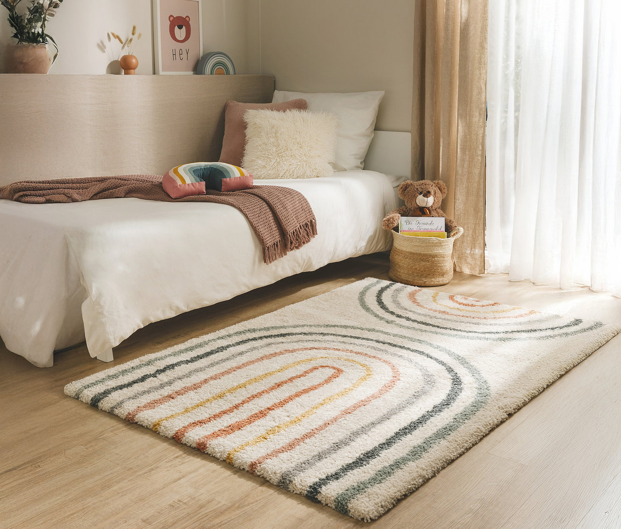 Ein Kinderzimmer mit einem Bett, einem Teppich mit Regenbogenmuster und einem Teddybären in einem Korb.