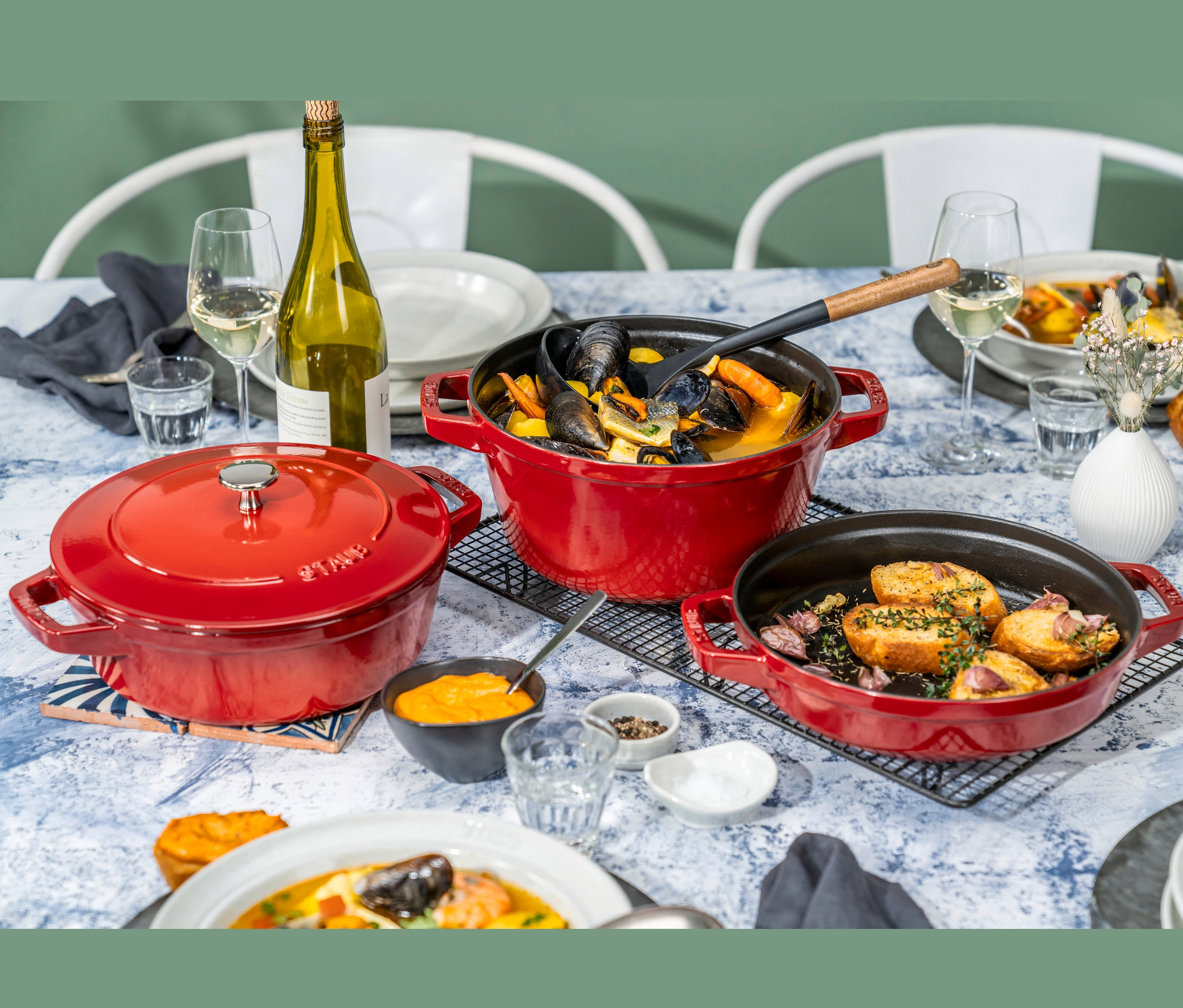 Auf einem Tisch stehen drei rote STAUB Kochtopf-Set »La Cocotte« Kochtöpfe und Speisen.