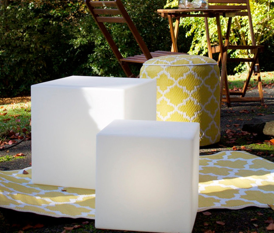 Zwei LED-Leuchtwürfel und ein gelber Hocker auf einer Decke im Freien, im Hintergrund ein Tisch mit Stühlen.