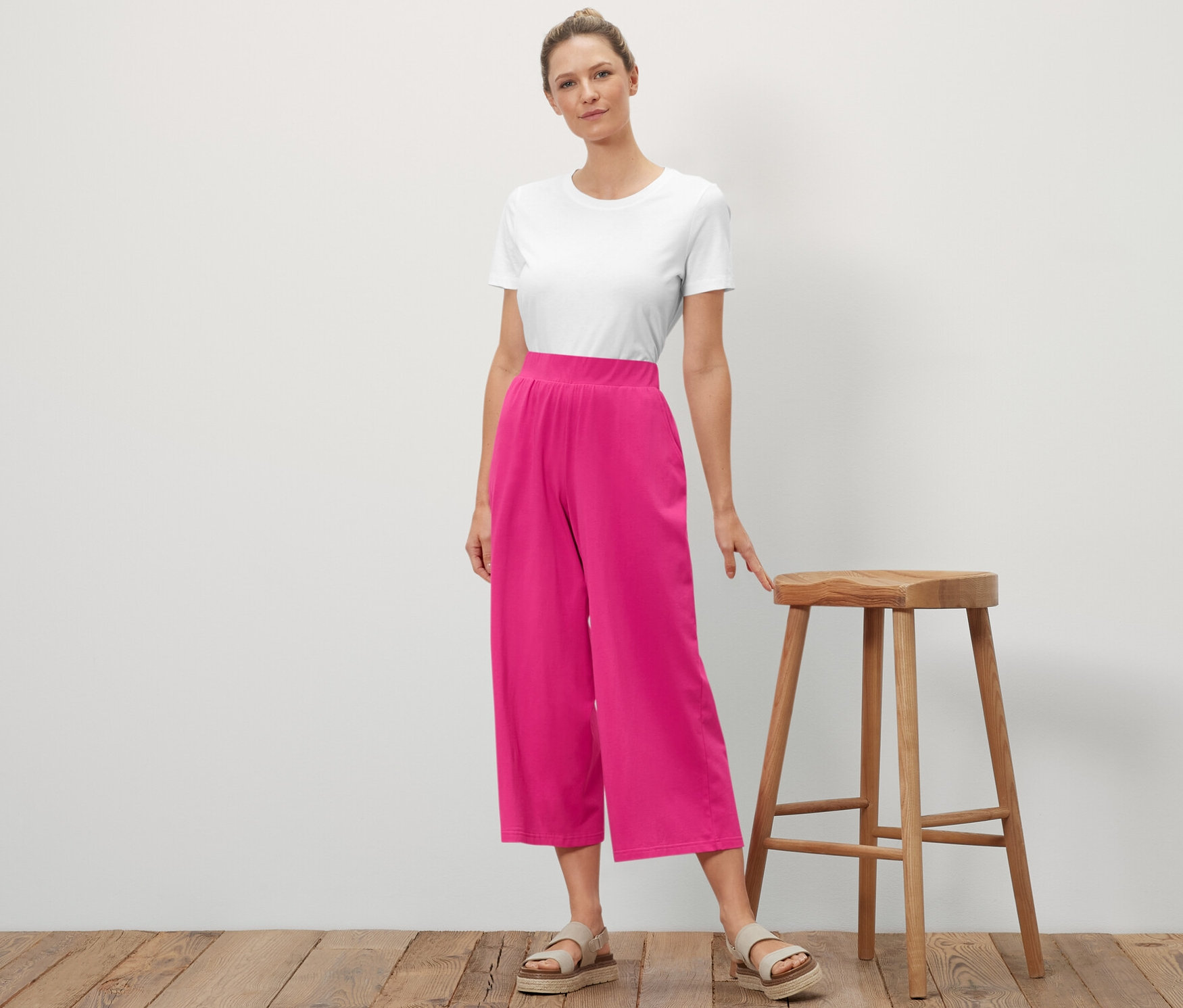 Eine Frau steht neben einem Holzhocker und posiert. Sie trägt ein weißes T-Shirt und eine rosafarbene Jersey-Culotte.