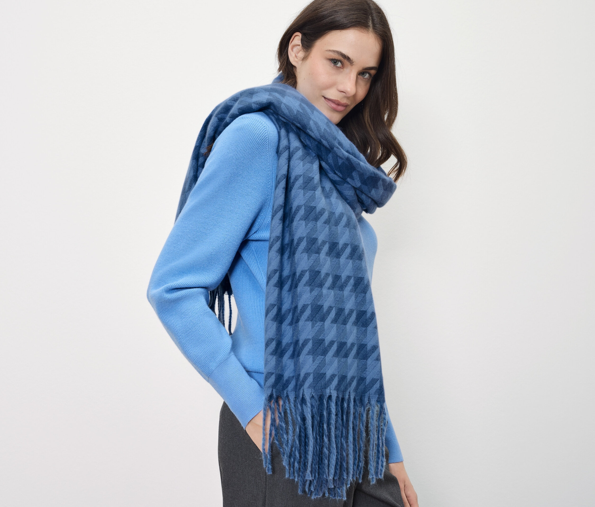 Eine Frau mit braunen Haaren trägt einen blauen Pullover und einen blauen Schal mit Karomuster.