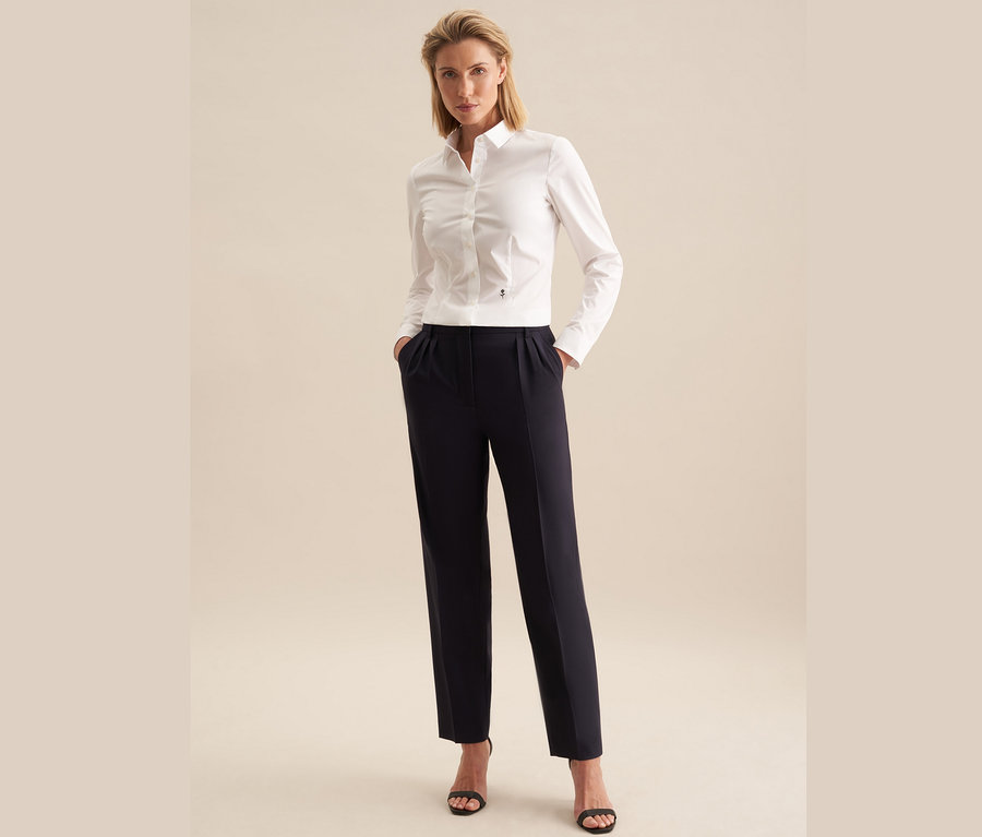 Frau steht mit Händen in den Hosentaschen. Sie trägt eine weiße Seidensticker Slimfit Bluse, dunkelblaue Hosen und schwarze Sandalen.