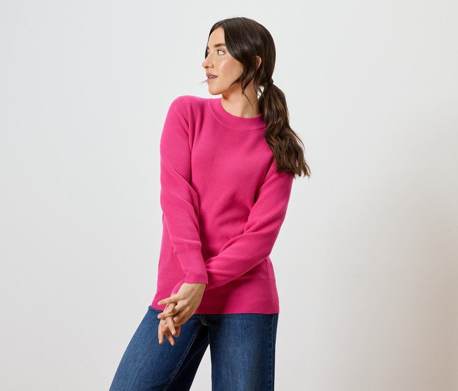 Frau mit braunen Haaren und blauen Augen posiert in einem pinkfarbenen Pullover und blauen Jeans.