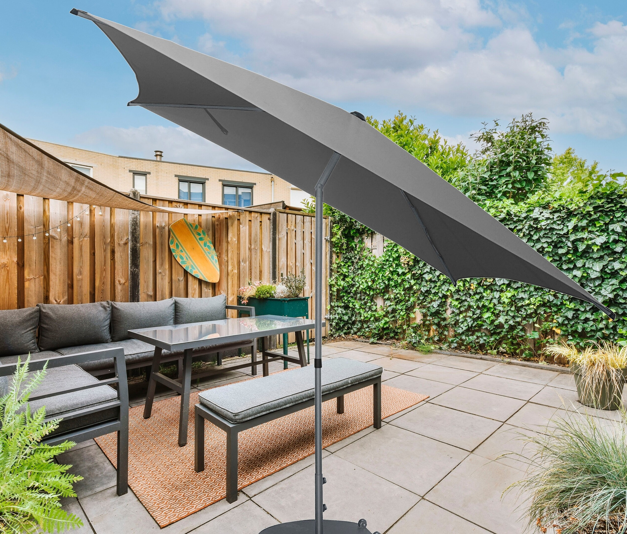 Grauer Leco Marktschirm 250 x 250 cm mit Bezug steht über einem grauen Sofa, Tisch und Bank auf einer Terrasse.