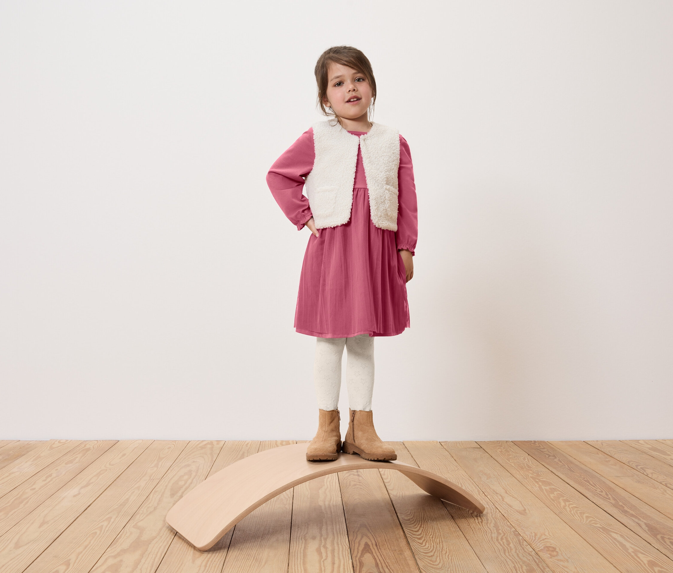 Mädchen steht auf einem hölzernen Balance Board, trägt ein rosa Kleid, eine weiße Weste und beige Stiefel.