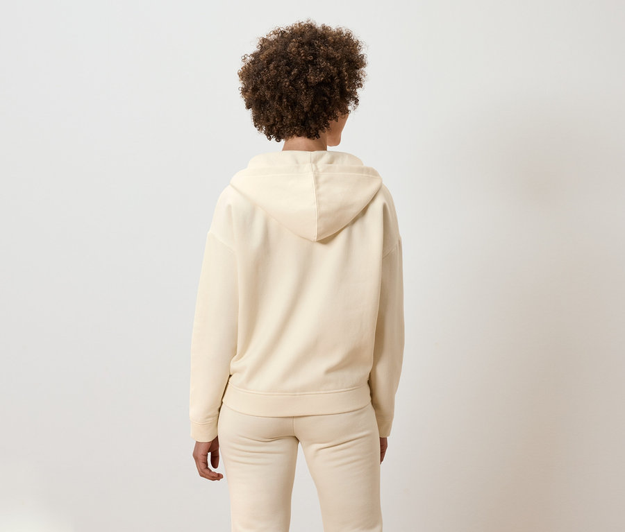 Rückansicht einer Frau mit lockigem Haar, die ein beigefarbenes Kapuzen-Outfit trägt.
