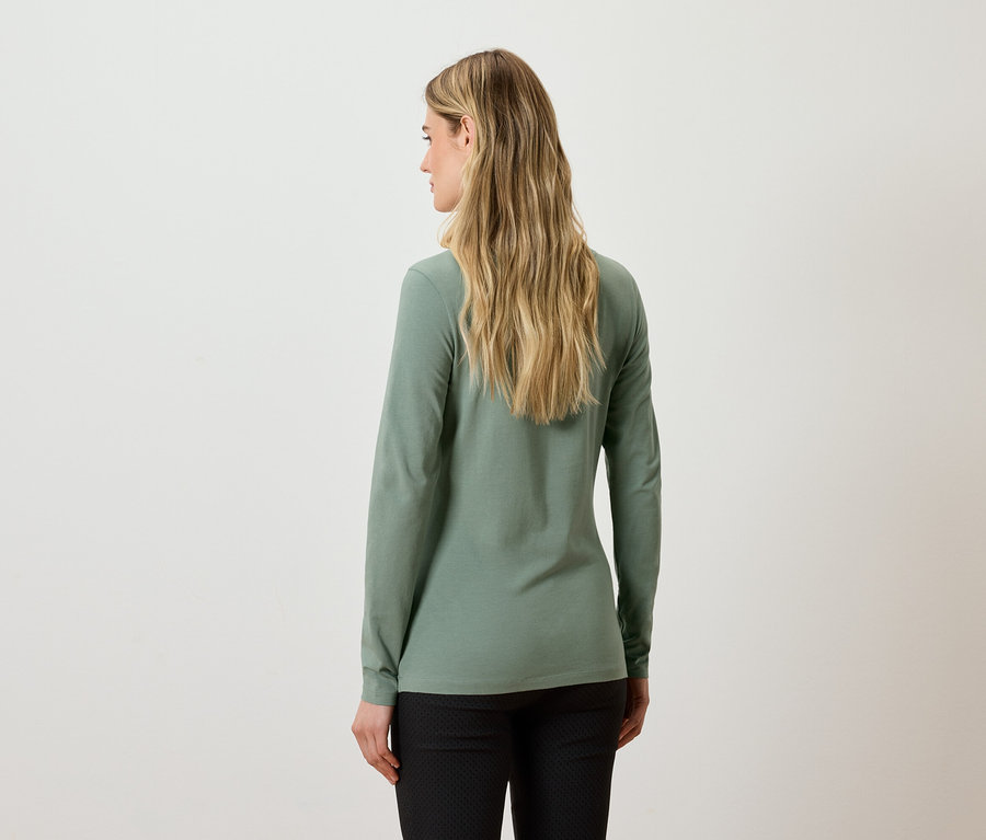 Rückansicht einer Frau mit langen blonden Haaren in einem grünen Langarmshirt und einer schwarzen Hose.