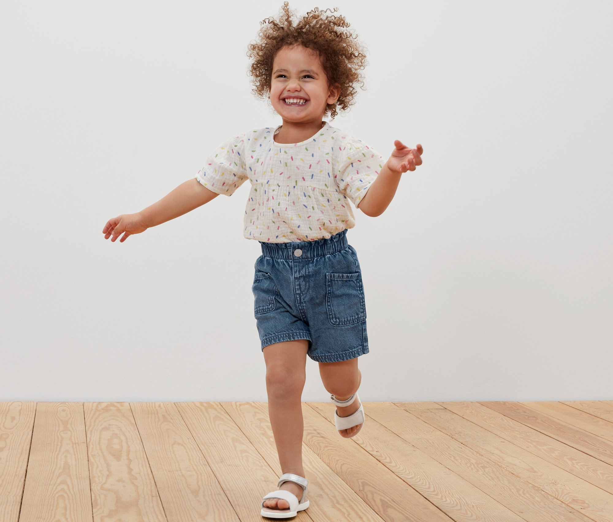 Ein lächelndes kleines Mädchen rennt über einen Holzboden und trägt Kinder-Jeansshorts und Sandalen.