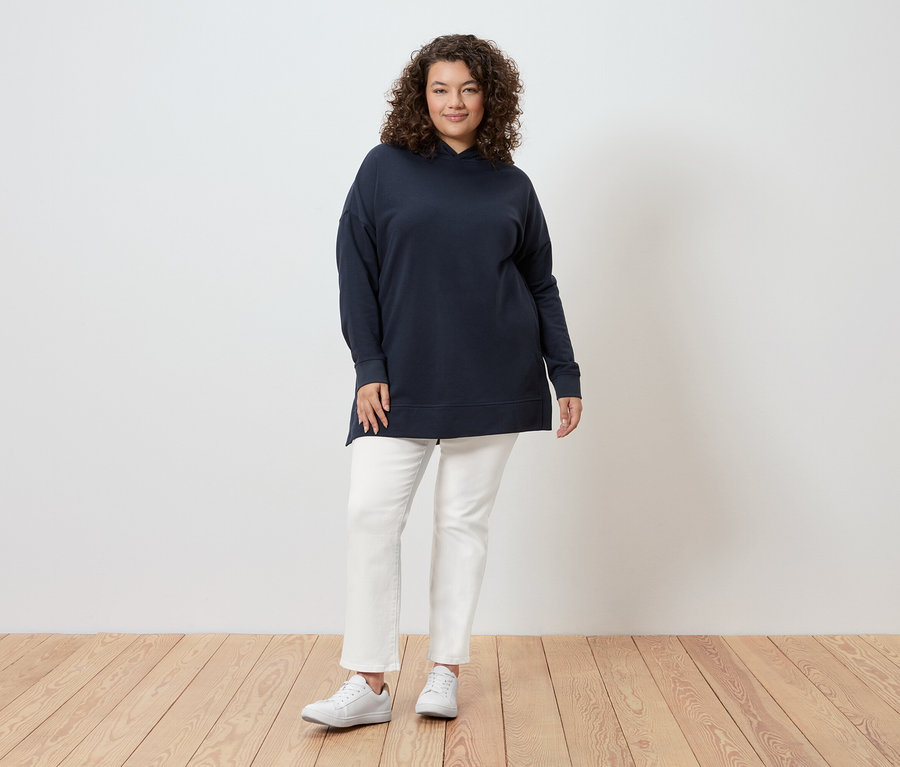 Eine Frau mit lockigem Haar steht vor einer Wand und trägt einen marineblauen Kapuzenpullover, weiße Jeans und weiße Turnschuhe.