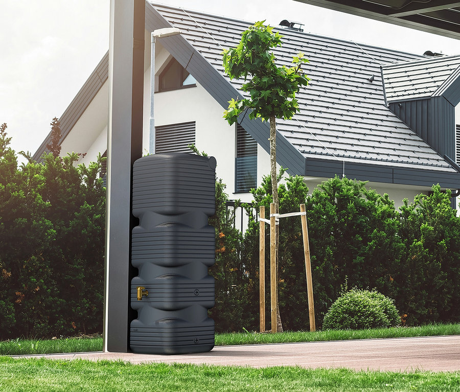 Eine graue Regenwassertonne steht neben einem modernen Haus mit Garten.