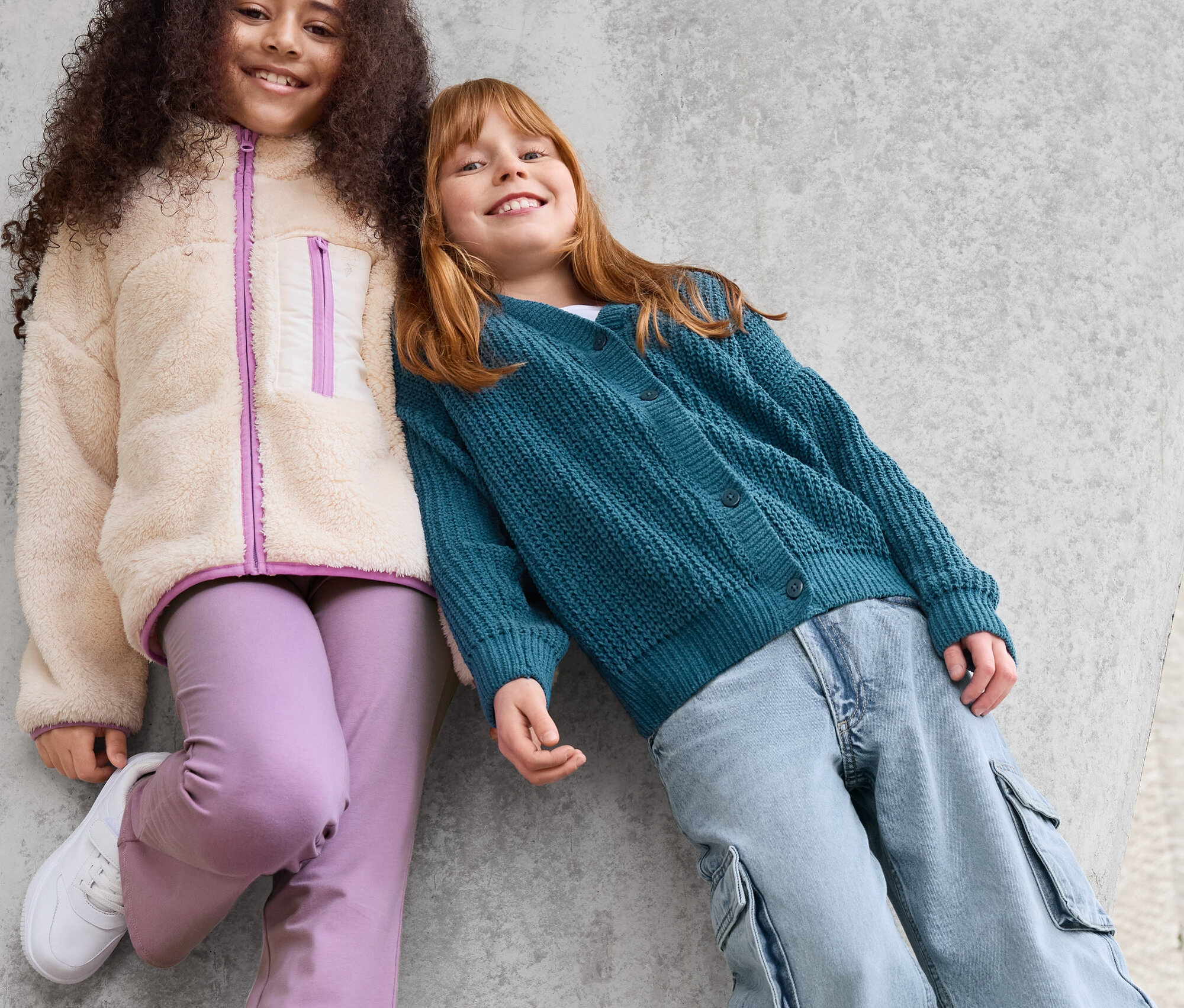 Zwei Mädchen lehnen an einer Betonwand. Das Mädchen links trägt eine Jacke aus Teddyfell, eine lila Hose und weiße Turnschuhe. Das Mädchen rechts trägt eine grüne Strickjacke und eine blaue Hose.