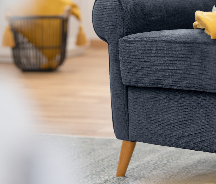 Detailaufnahme eines dunkelblauen Ohrensessels "Anton" mit Holzbeinen und gelber Decke. Im Hintergrund ein Weidenkorb mit gelbem Stoff.
