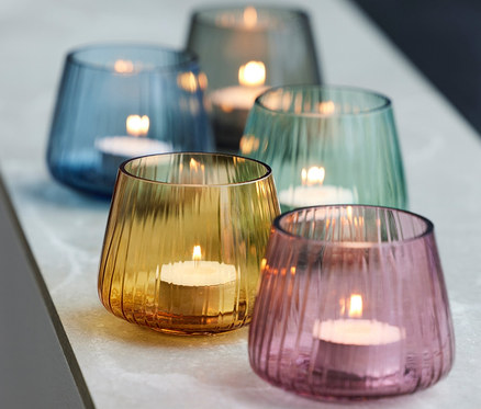 Fünf Teelichthalter »Kusintha« von Bitz aus Glas in Blau, Grün, Rosa und Gelb mit brennenden Teelichtern, die in einer Reihe auf einer hellen Oberfläche angeordnet sind.