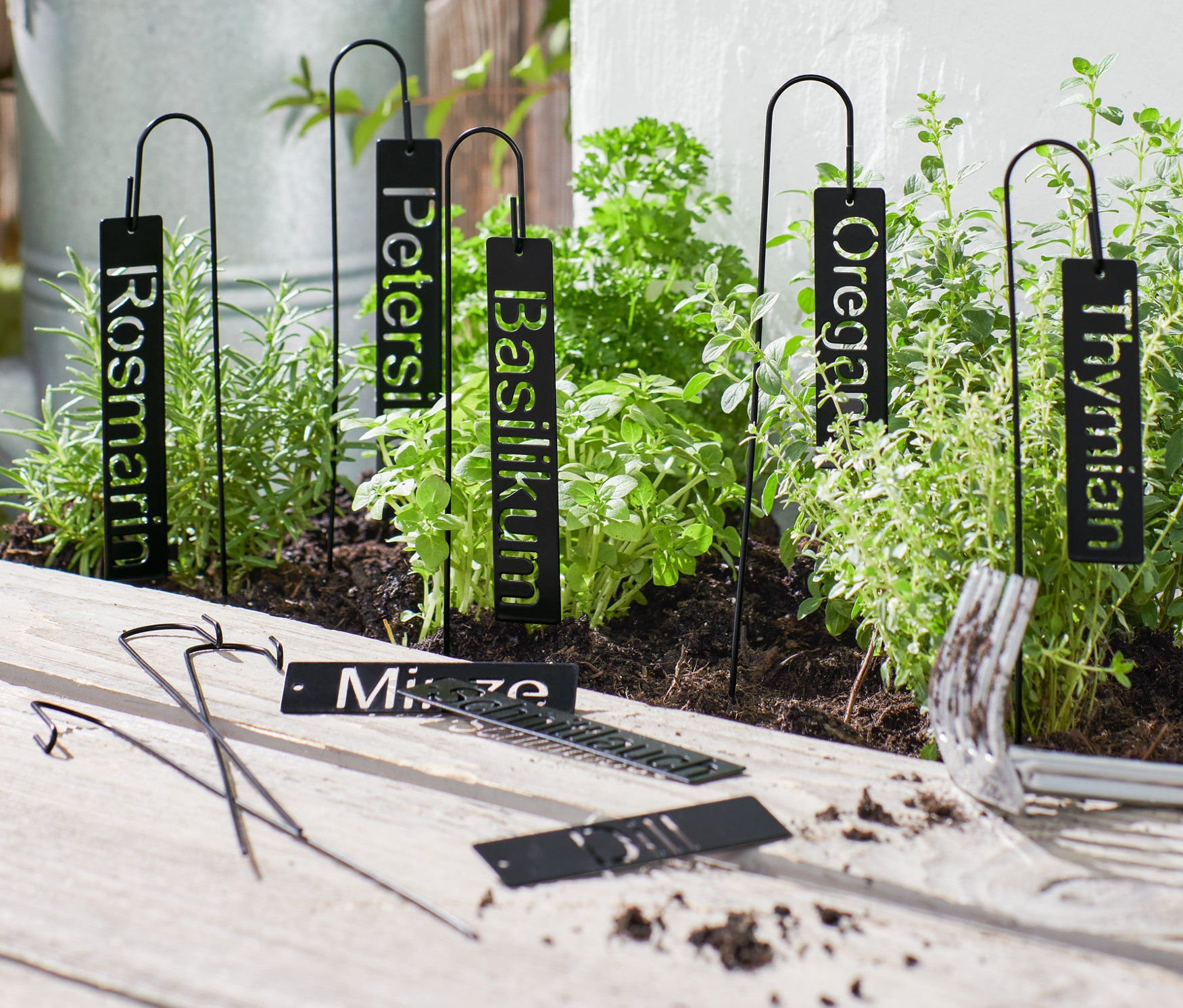 Schwarze Kräuterschilder stecken in einem Kräuterbeet, Gartengeräte liegen auf einem Holztisch.