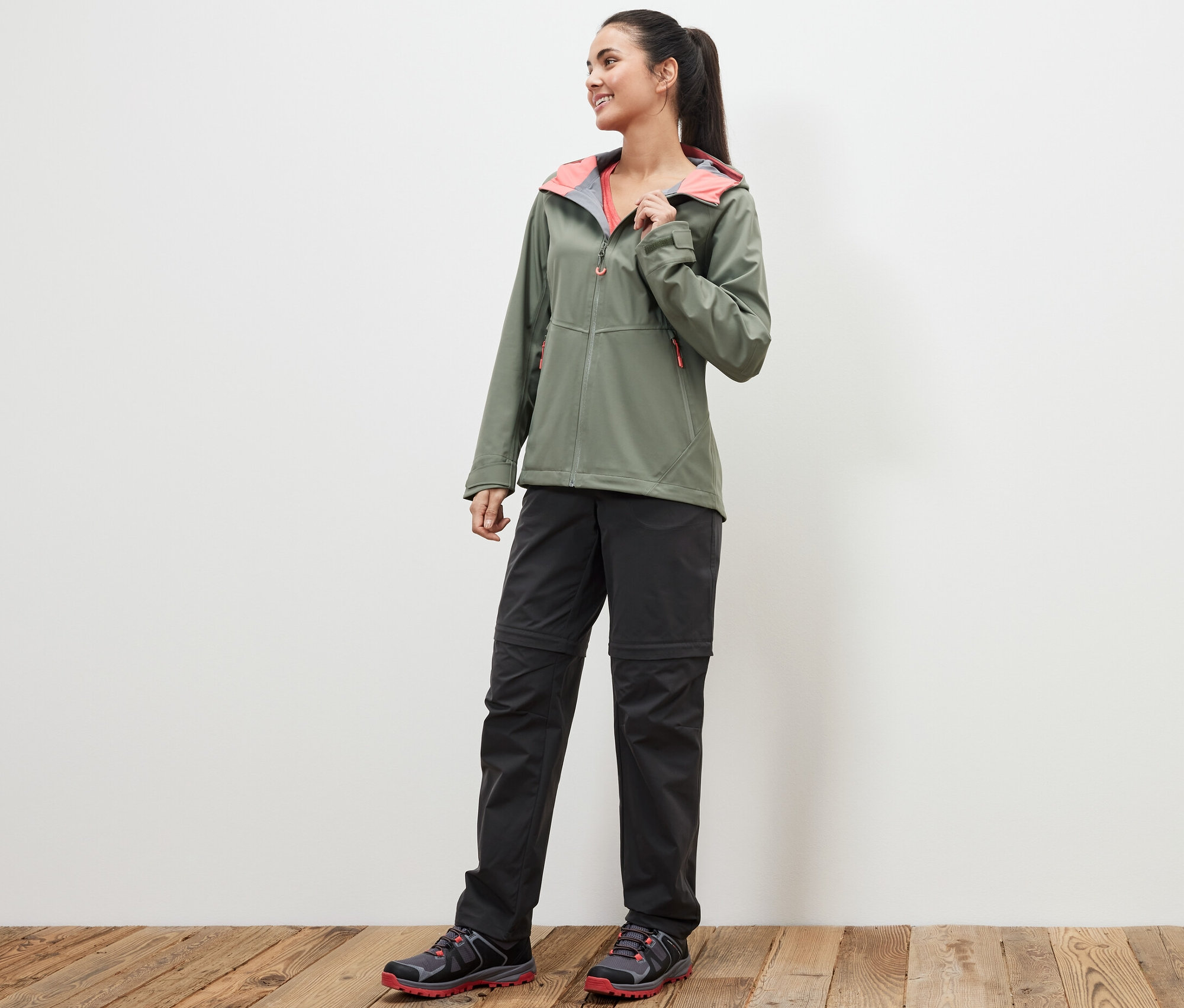 Eine Frau steht auf einem Holzboden und trägt eine grüne Jacke und eine schwarze 2-in-1-Funktionshose.