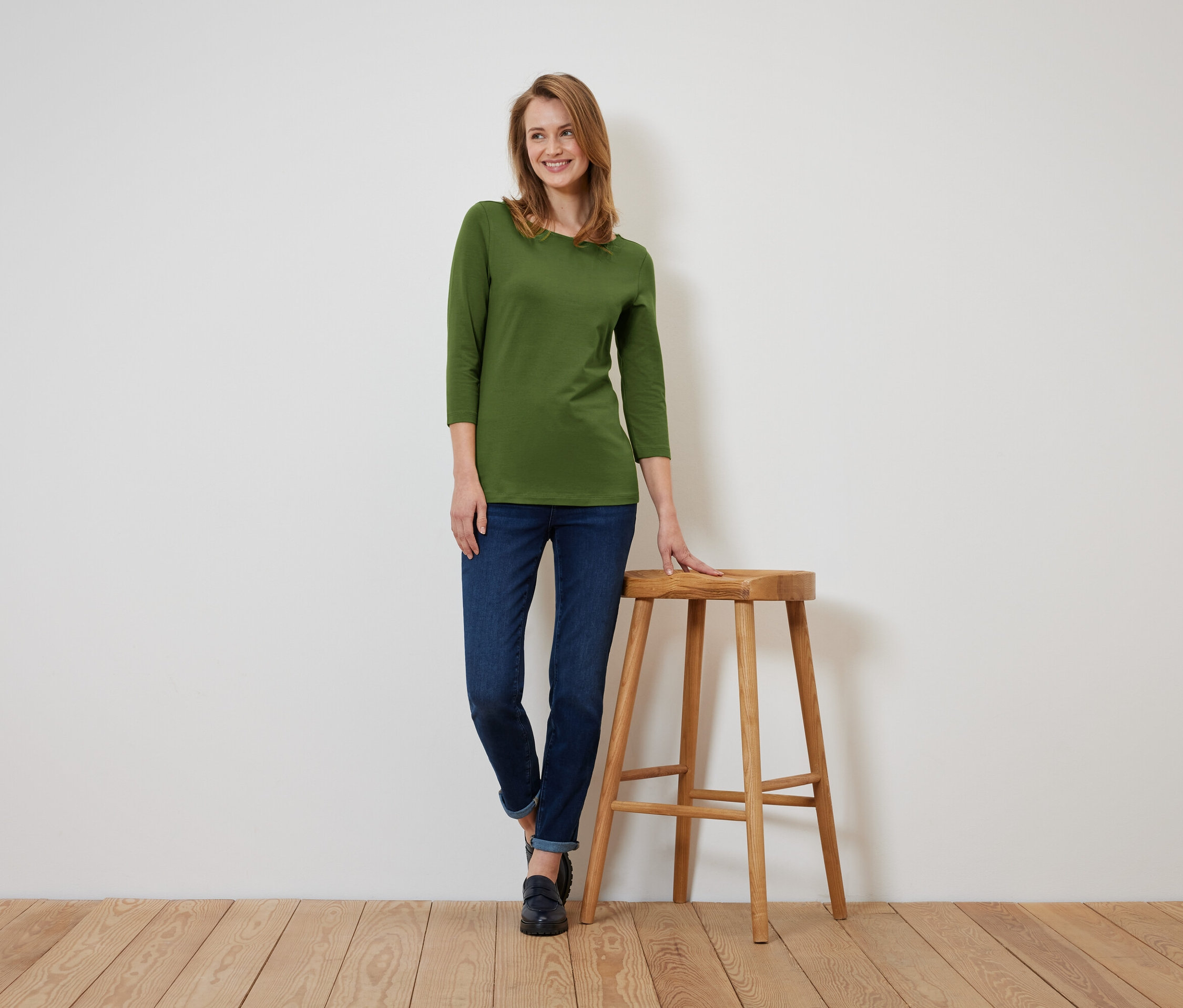 Eine Frau in einem olivgrünen Dreiviertelarm-Shirt und dunkelblauen Jeans steht und lehnt sich an einen Holzhocker.