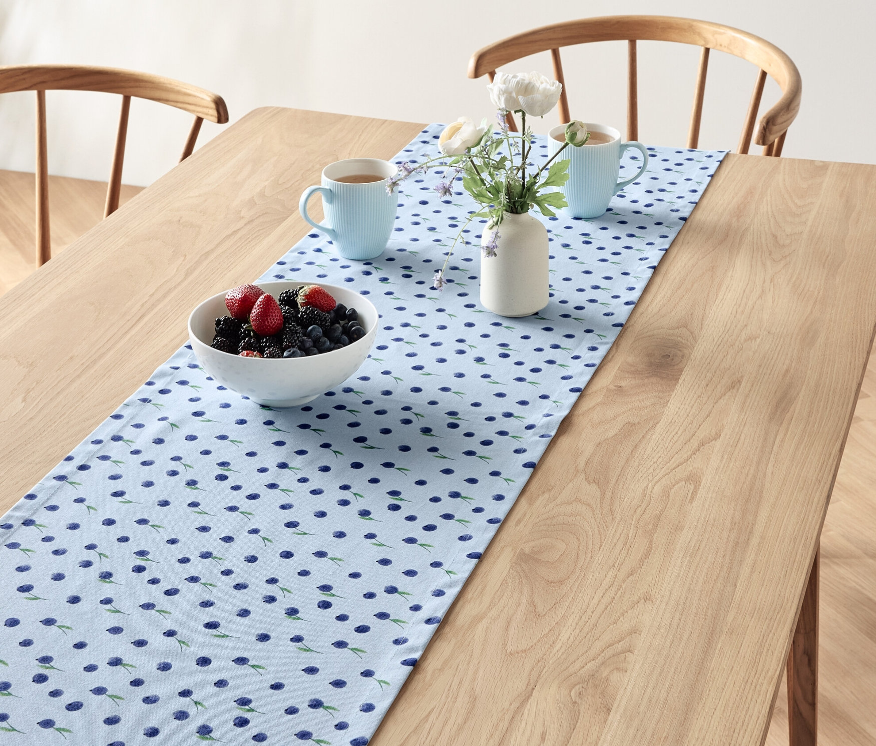 Auf einem Holztisch liegt ein Tischläufer mit Blaubeermuster, darauf eine Schale mit Beeren, zwei blaue Tassen und eine Vase mit Blumen.