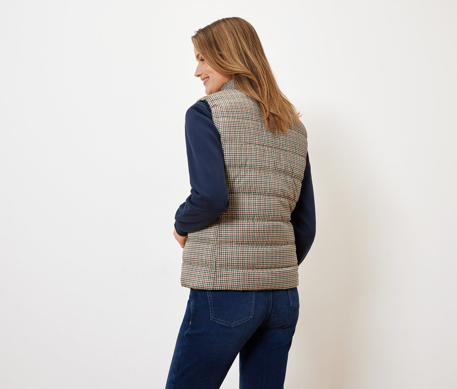 Frau mit dem Rücken zur Kamera trägt eine Steppweste mit Hahnentrittmuster, einen blauen Pullover und Jeans.