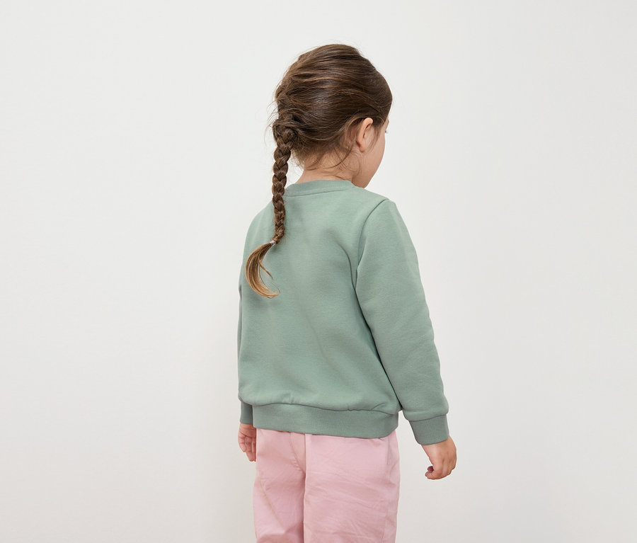 Rückansicht eines Mädchens mit Zopf, bekleidet mit einem grünen Pullover und einer rosa Hose.