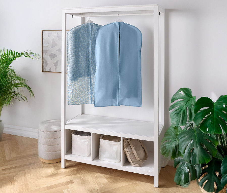 Zwei Kleidersäcke hängen an einer weißen Garderobe, mit Regalen und Fächern darunter. Eine Zimmerpflanze steht links und rechts.