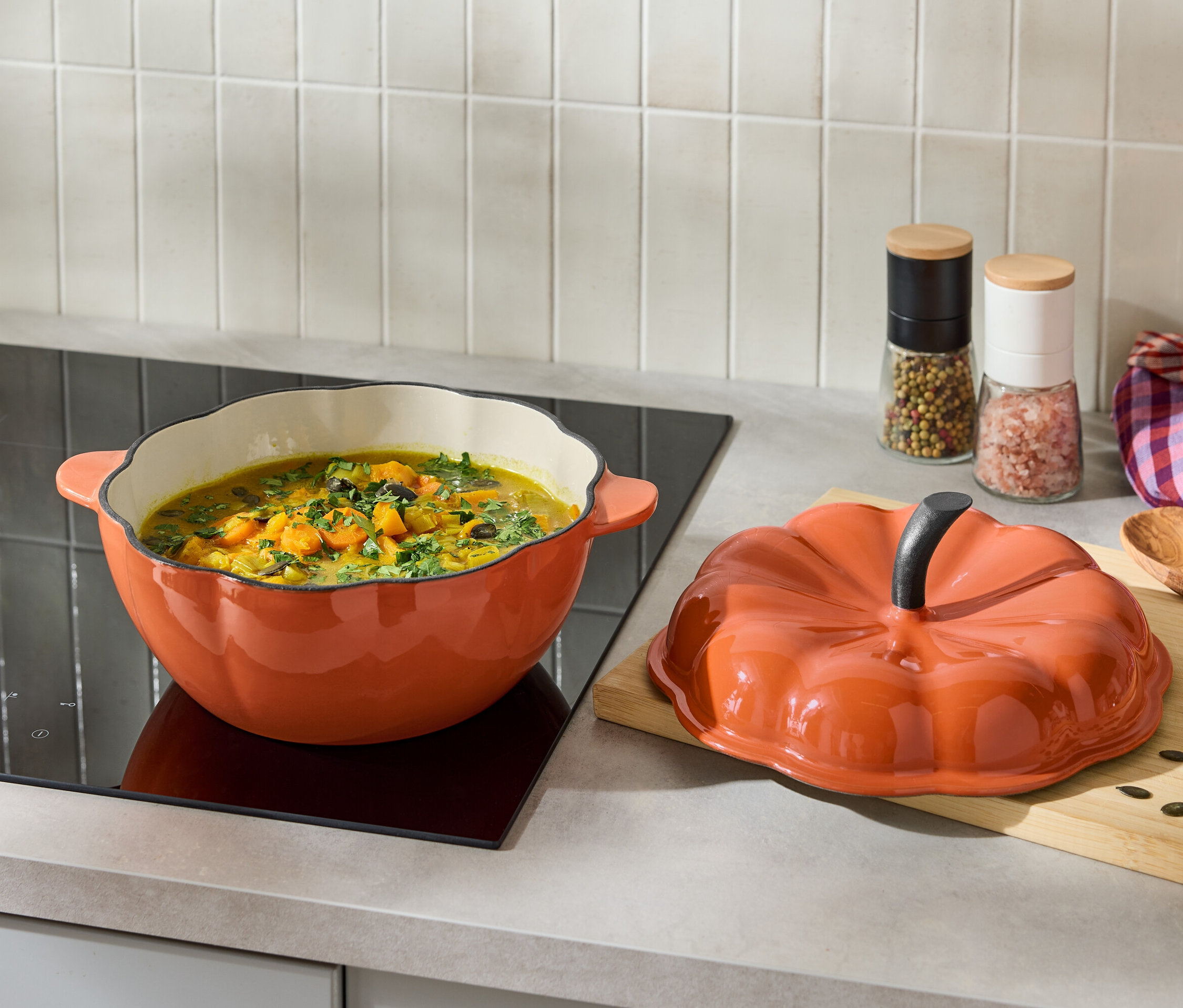 Orangefarbener Topf mit Suppe auf einem Induktionskochfeld neben einem orangefarbenen Deckel in Kürbisform.