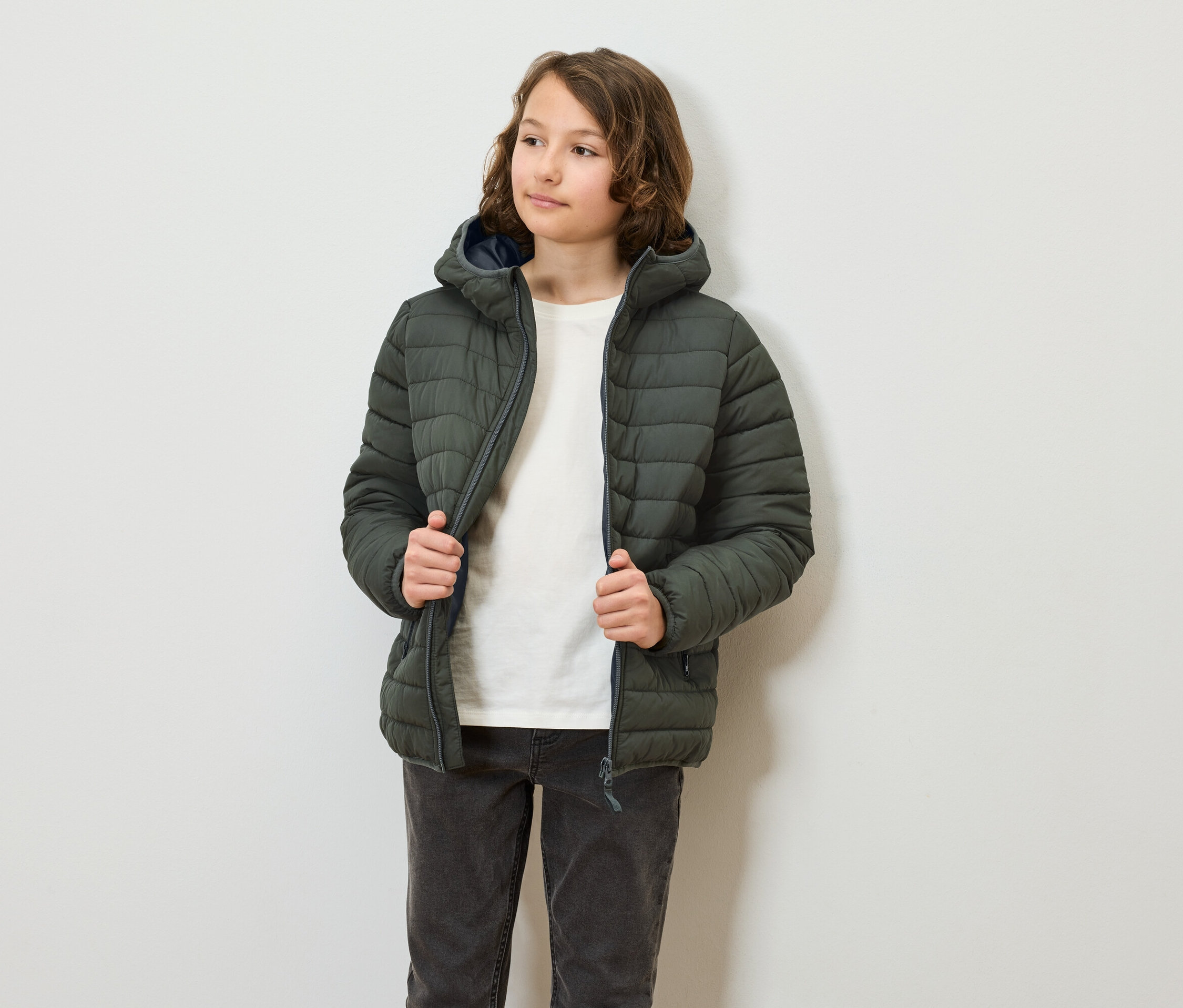 Eine jugendliche Person trägt eine olivgrüne Steppjacke mit Kapuze und schliesst sie. Die Person trägt Jeans und ein T-Shirt.