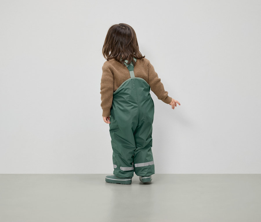 Ein Kind mit braunem Pullover und grüner Latzhose steht mit dem Rücken zur Kamera.