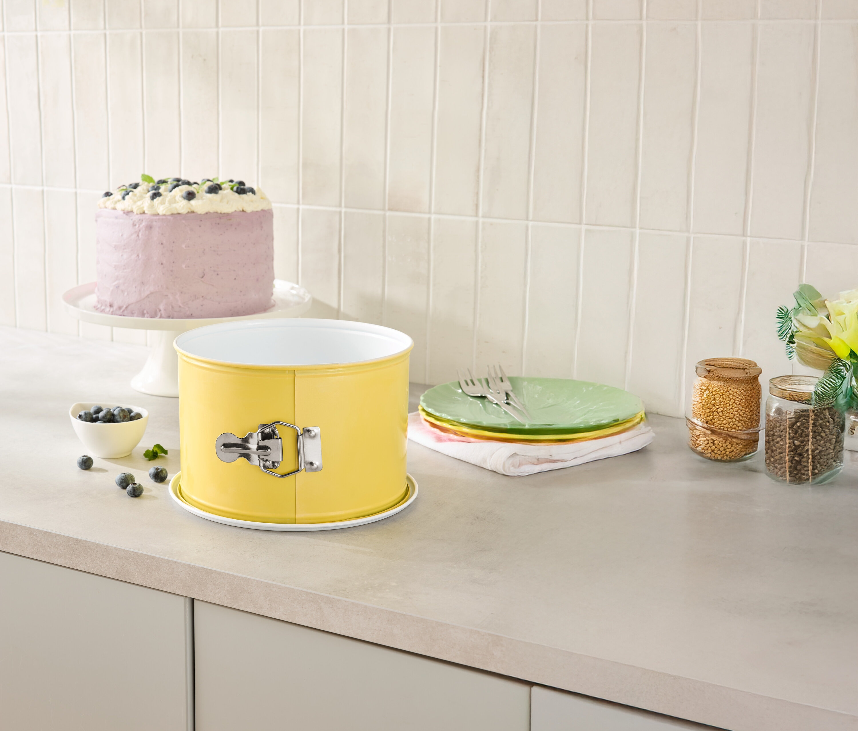 Auf einer Küchentheke stehen eine gelbe Springform, ein Kuchen auf einem Tortenständer, Teller und Vorratsgläser.