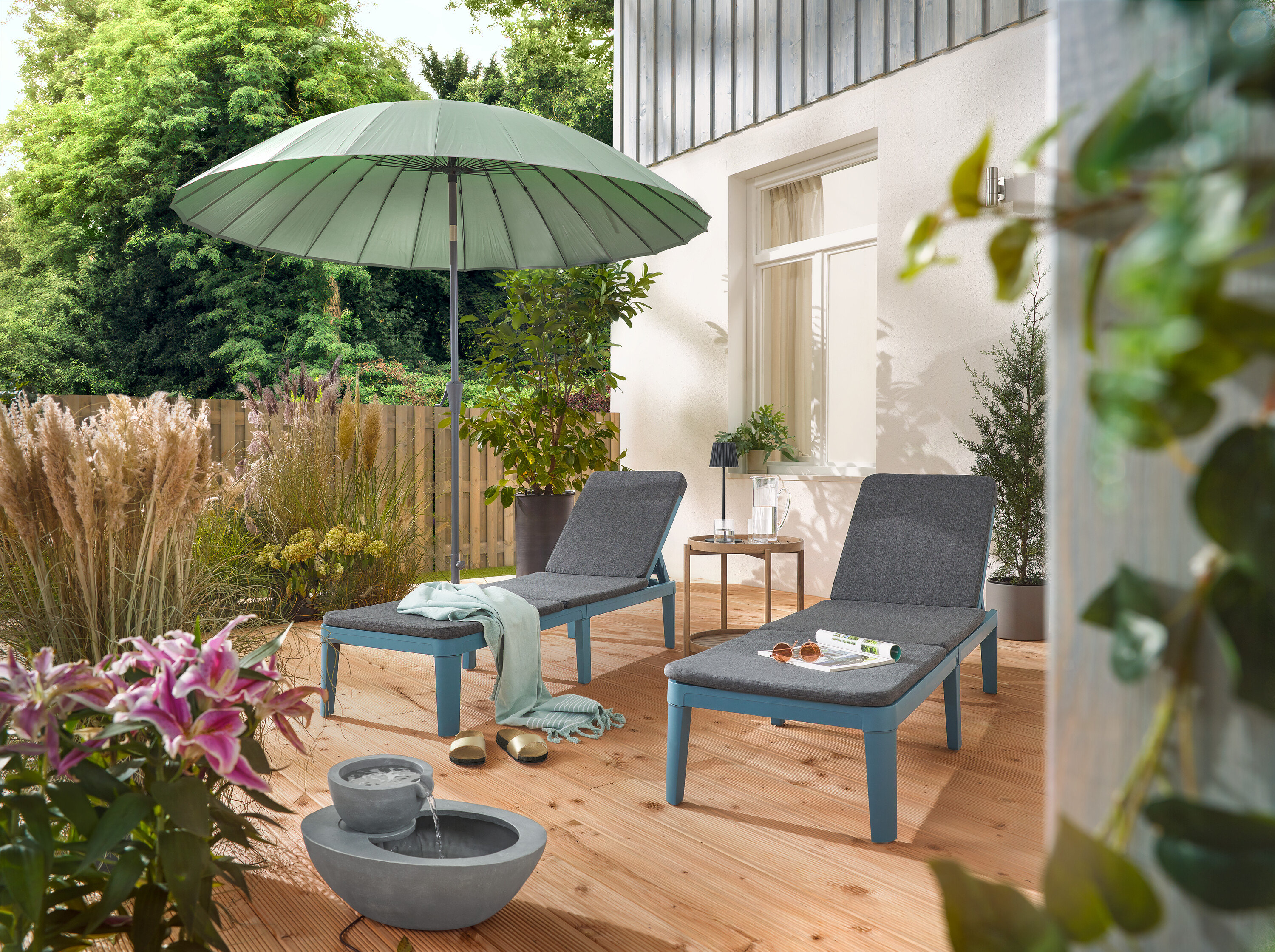 Auf einer Terrasse stehen zwei Liegen unter einem hellgrauen Leco Shanghai Sonnenschirm und neben einem Solar-Gartenbrunnen.
